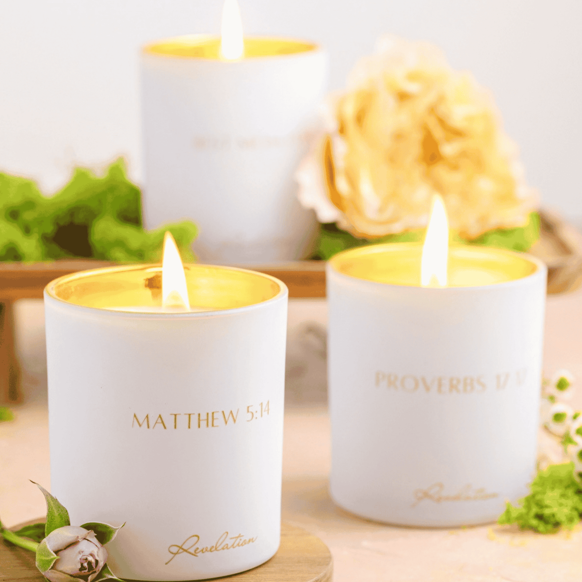 Three lit Revelation Candles with gold interiors, surrounded by pale roses and greenery.